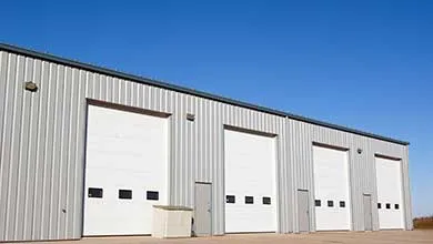 Gray corrugated metal building with three white roll-up garage doors, under a clear blue sky.