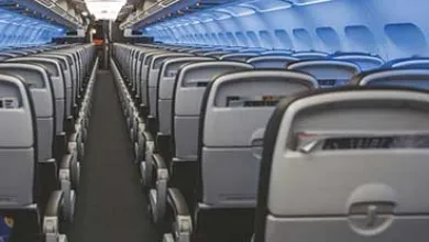 Empty airplane cabin interior: gray seats, white overhead lights, blue window lighting.