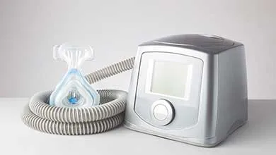 Silver CPAP machine with digital screen, gray coiled hose, and clear/blue nasal mask on a white surface.