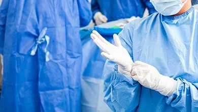 A medical professional in a blue surgical gown puts on white sterile gloves in an operating room.