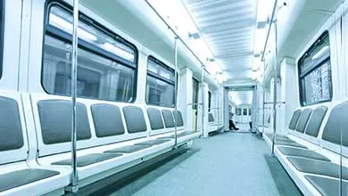 Empty modern subway car with rows of grey seats along both sides of a central aisle, lit by cool blue-white lighting.