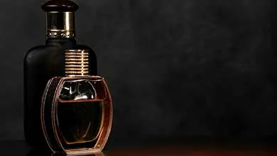 Two perfume bottles, one clear glass with gold accents and one matte black, sit side by side on the left against a dark textured background.