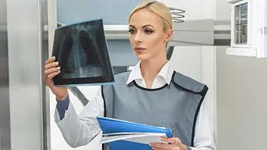 A female doctor wearing a lead apron over a white coat examines a chest X-ray.
