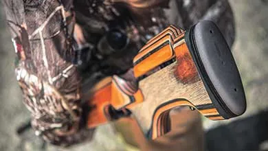 Overhead close-up of a laminated wood rifle stock with orange and black stripes, held by a person wearing camouflage.