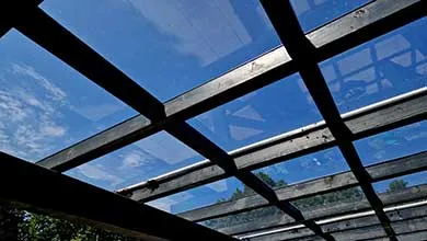 Low-angle view up through a glass roof with dark beams, showing a bright blue sky and white clouds.