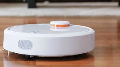 A white, round robot vacuum with an orange sensor on top sits on a hardwood floor.
