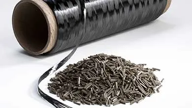A large spool of black carbon fiber sits on a white surface behind a pile of small, chopped carbon fiber pellets.