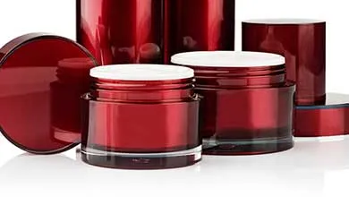 Glossy red cosmetic containers on a reflective white surface, with two open jars showing white inner lids in front and taller cylindrical bottles behind them.