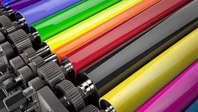Close-up of large, glossy rollers in purple, blue, green, yellow, red, black, and magenta, arranged diagonally on a printing press, with gears visible on the left.