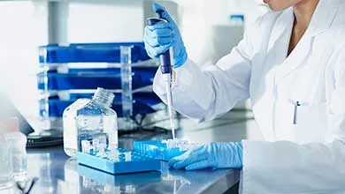 A scientist in a white lab coat and blue gloves uses a pipette to transfer liquid into test tubes in a blue rack on a lab bench.