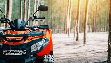 Orange all-terrain vehicle parked on a dirt trail in a forested outdoor setting with sunlight filtering through trees.
