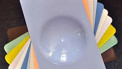 A light blue rectangular sample with a raised, glossy circular dome rests on top of fanned color swatches in yellow, green, brown, white, and orange, set against a dark background.