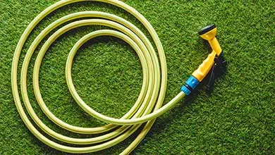 A coiled pale yellow garden hose with a yellow and blue spray nozzle rests on green artificial grass.