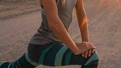 A woman in a gray tank top and teal-and-black striped leggings performs a low lunge stretch on a dirt path, hands resting on her front knee. Warm golden-hour sunlight creates a lens flare in the background.